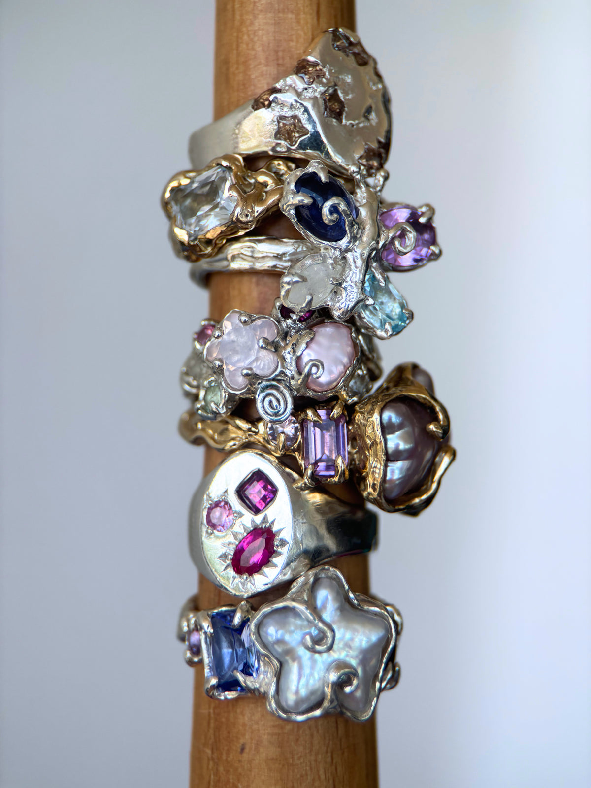 A stack of nine unique vintage-style rings displayed on a wooden ring stand against a light gray background. The rings feature various textured metal bands in silver, gold, and bronze, adorned with colorful gemstones including pink, purple, blue, and clear crystals.